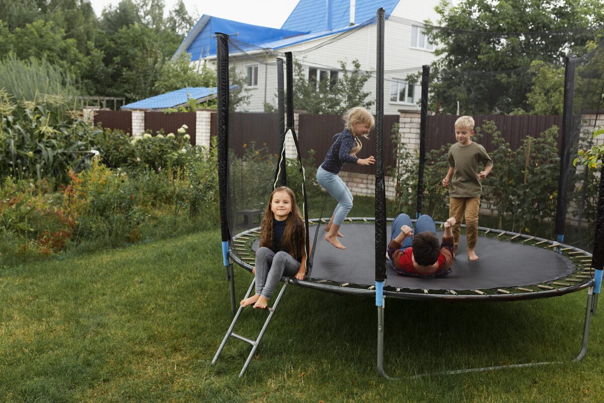 Four children enjoy a high bounce trampoline in a lush backyard, safely enclosed with a net and surrounded by greenery and a residential home.