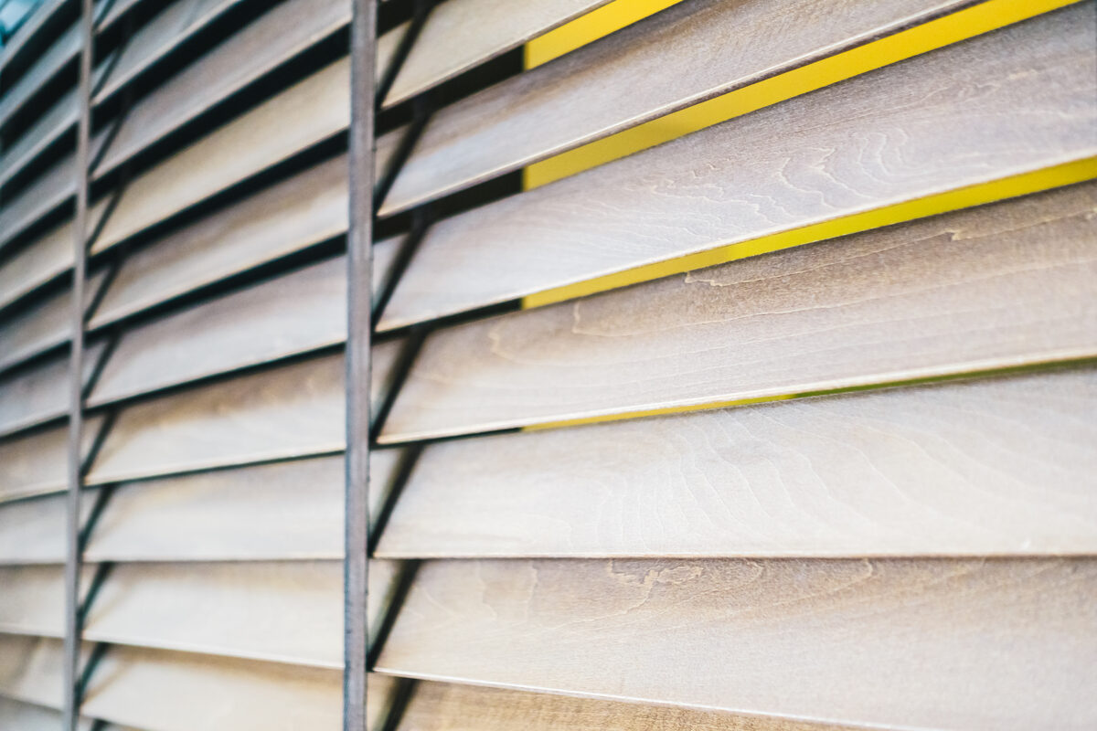 Close-up of wooden sunscreen roller blinds partially open, filtering warm light through horizontal slats with a vibrant yellow backdrop.